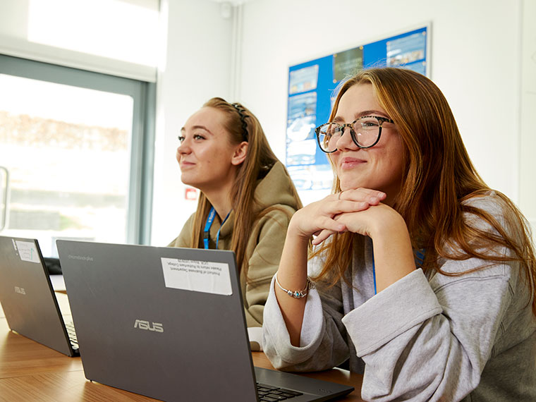 Two students sat side by side at a desk with their laptops open
