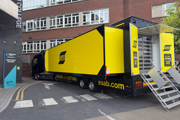 A large yellow and black lorry parked next to a building, the back of the lorry is open and stairs go from the lorry to the floor