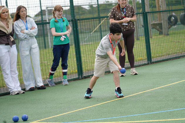Someone wearing beige shorts and a white top about to throw a small blue bowling ball, four people stood behind watcching