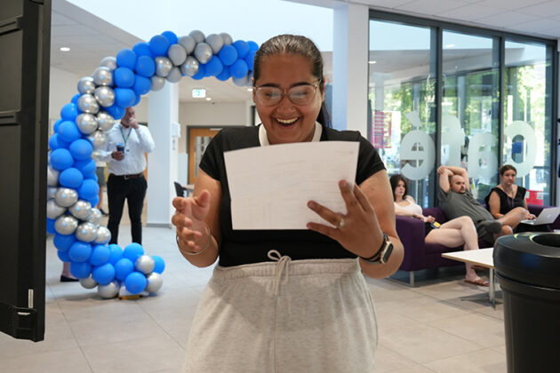A female looking happy looking down at a piece of paper in her hands, a balloon arch in the background