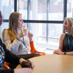 Three females sat around a table smiling and talking, one holding a drinking cup