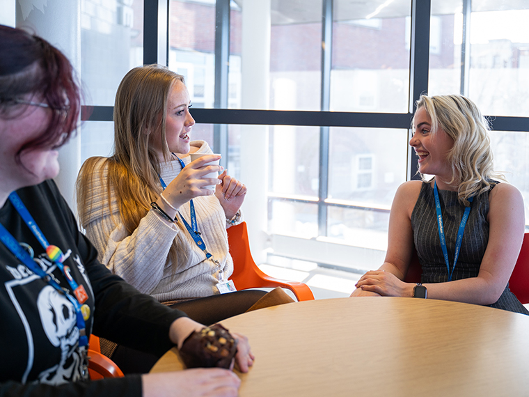 Three females sat around a table smiling and talking, one holding a drinking cup